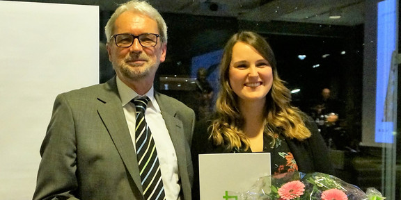 Bild von Dr. Heinrich Tröster und Teresa Mann. Mann in grauem Anzug mit kurzen weißen Haaren und einem kurzen weißem Bart und Brille ist links im Bild und lächelt. Daneben ist eine lächelnde Frau mit einem schwarzen Kleid mit rosa Blumen. Sie hat lange braune gewellte Haare und hält einen Blumenstrauß in einer und ein Dokument in der anderen Hand.