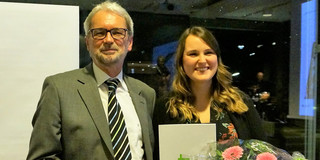 Lotte Kaliski Preis 2018 Bild von Dr. Heinrich Tröster und Teresa Mann. Mann in grauem Anzug mit kurzen weißen Haaren und einem kurzen weißem Bart und Brille ist links im Bild und lächelt. Daneben ist eine lächelnde Frau mit einem schwarzen Kleid mit rosa Blumen. Sie hat lange braune gewellte Haare und hält einen Blumenstrauß in einer und ein Dokument in der anderen Hand.