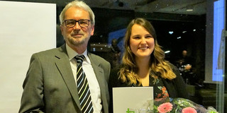 Lotte Kaliski Preis 2018 Bild von Dr. Heinrich Tröster und Teresa Mann. Mann in grauem Anzug mit kurzen weißen Haaren und einem kurzen weißem Bart und Brille ist links im Bild und lächelt. Daneben ist eine lächelnde Frau mit einem schwarzen Kleid mit rosa Blumen. Sie hat lange braune gewellte Haare und hält einen Blumenstrauß in einer und ein Dokument in der anderen Hand.