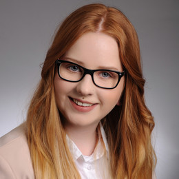 Proträt von Jil Brambrink. Frau mit langen dunkelorangenen Haaren, einer Brille, lächelt in die Kamera. Sie trägt einen Piercing mittig über der Oberlippe. 