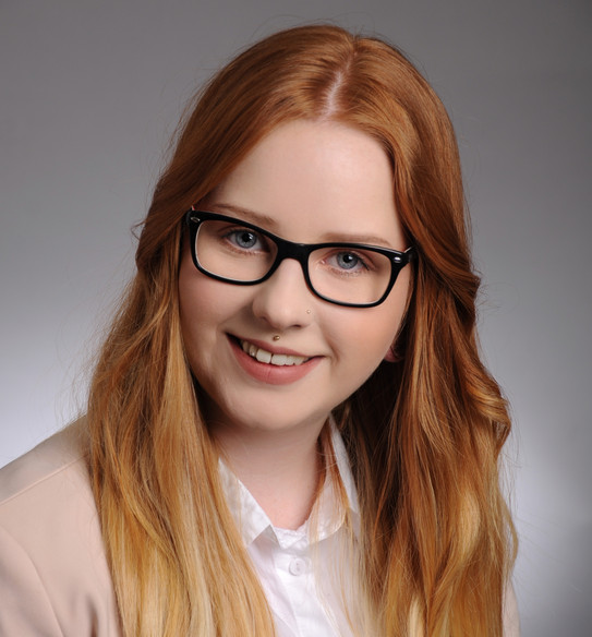 Jil Brambrink, M.A. (Wissenschaftliche Mitarbeiterin) Proträt von Jil Brambrink. Frau mit langen dunkelorangenen Haaren, einer Brille, lächelt in die Kamera. Sie trägt einen Piercing mittig über der Oberlippe.