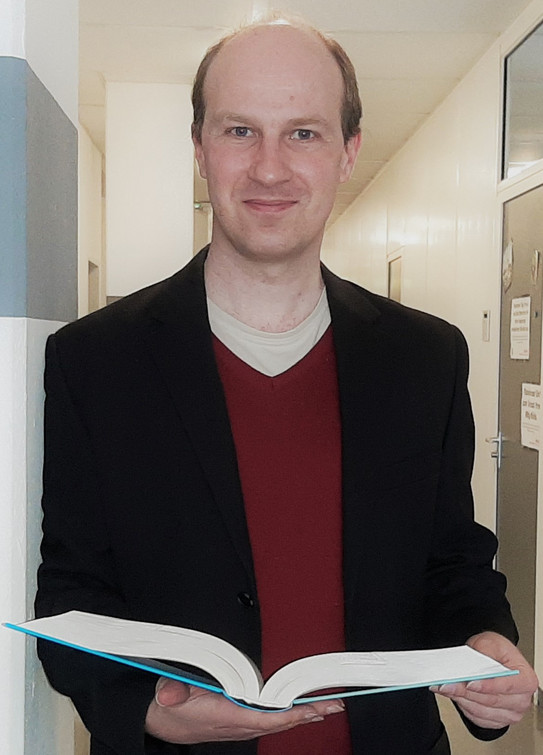 Portärt von PD Dr. Steffen Wild. Mann mit kurzen hellen Haaren lächelt in die Kamera. In den Händen hält er ein aufgeschlagenes Buch